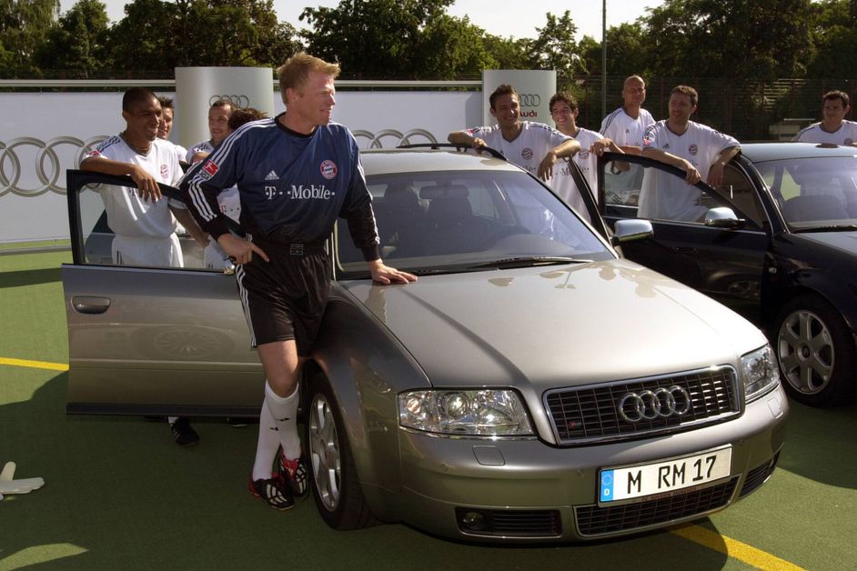 Oliver Kahn and other players of the 2002 FC Bayern squad stand around a silver Audi
