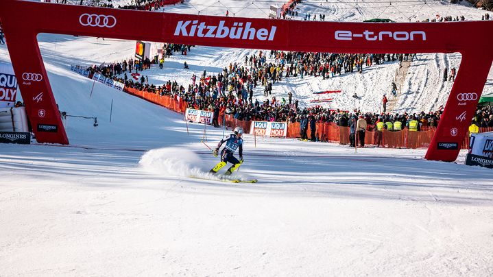 A skier braking at the finish line in Kitzbühel. An Audi logo and the words Kitzbühel and e-tron can be seen on the red finish arch.