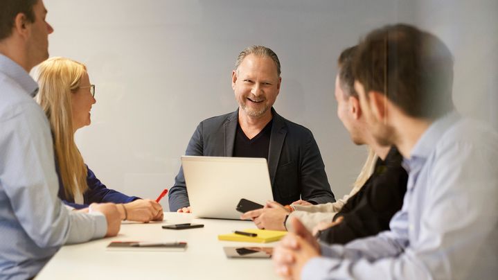 Mann sitzt an einem Meetingstisch mit seinen Angestellten und lacht