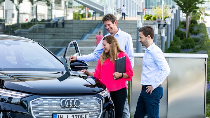 Three people are standing beside an open Audi car door, talking