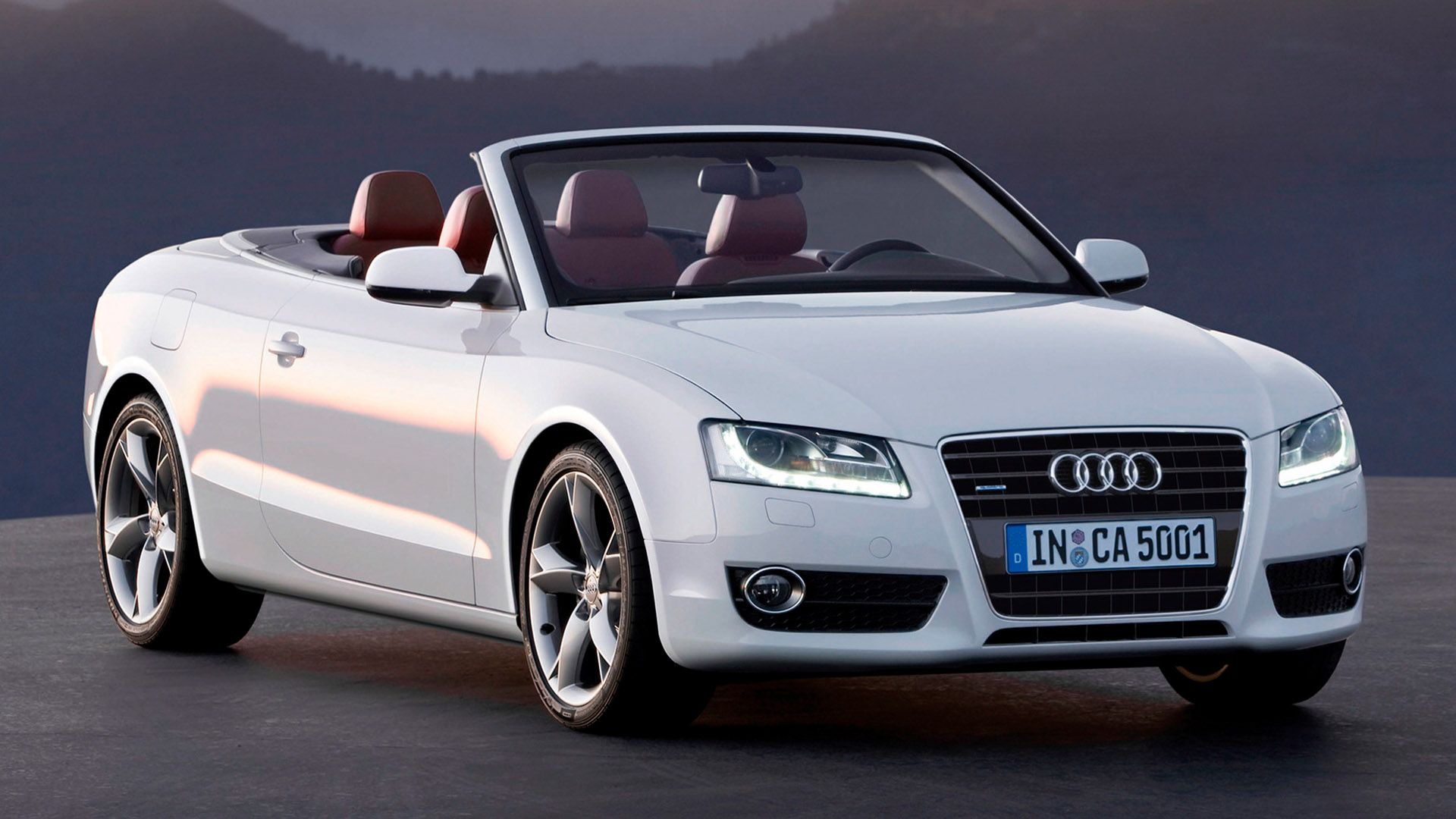White Audi A5 convertible with open top stands on a slope, forests can be seen in the background