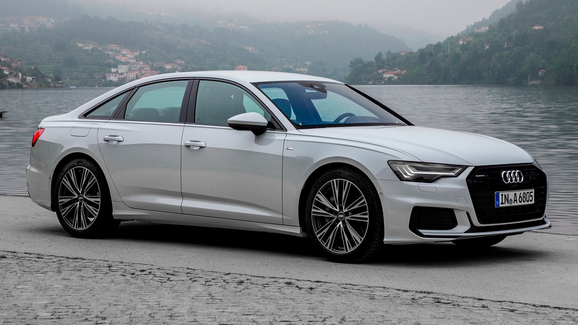 Light grey Audi A6 sedan standing in front of a lake, behind the lake is fog and mountains