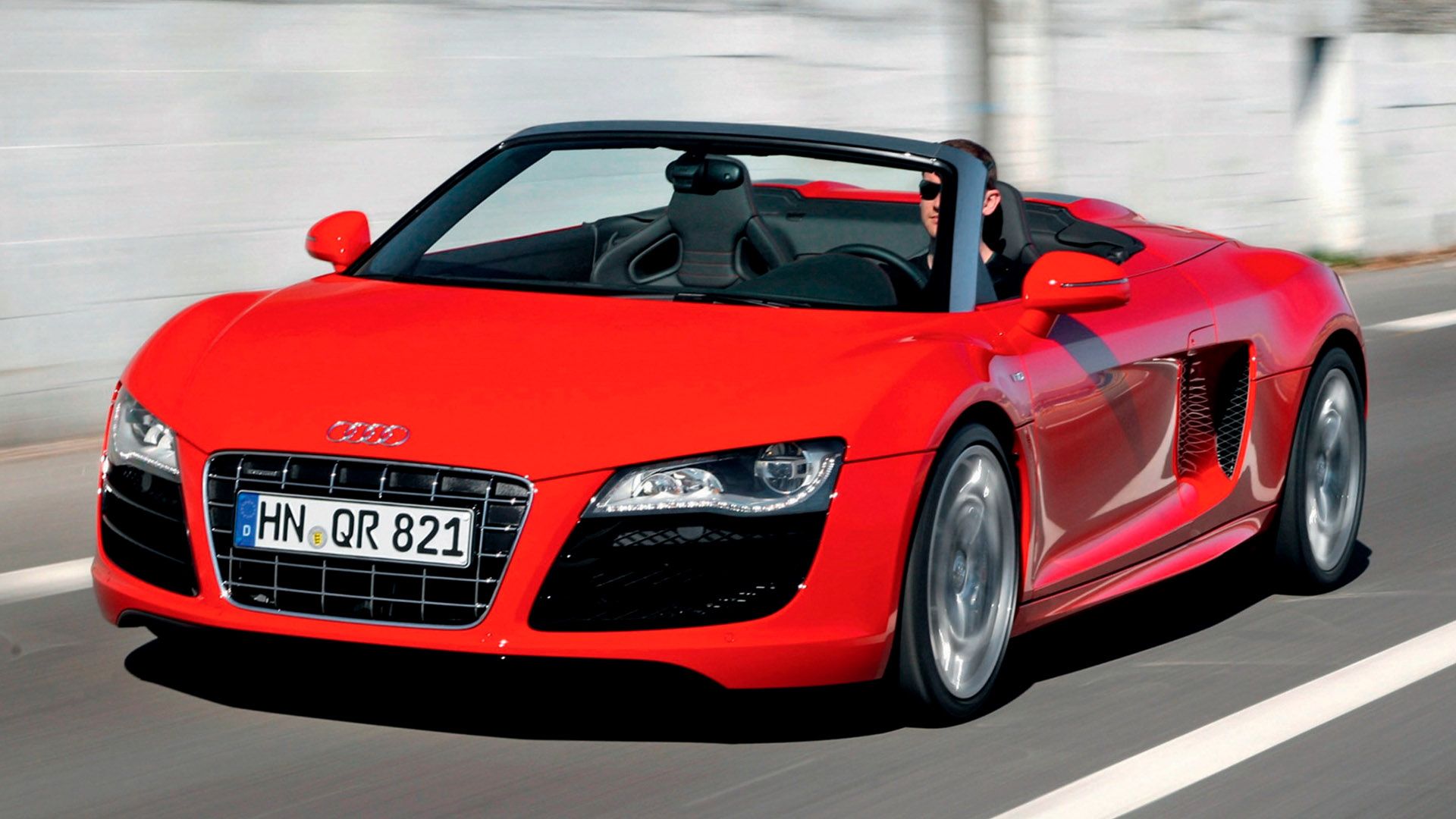 Red Audi R8 Spyder with open top travelling on a road, a man with sunglasses at the wheel