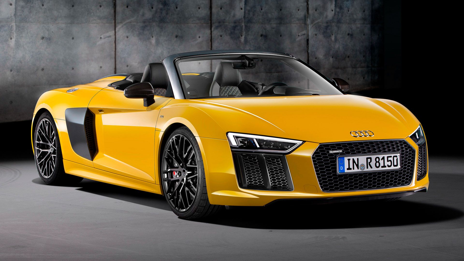 Yellow Audi R8 Spyder with open top in front of dark concrete wall