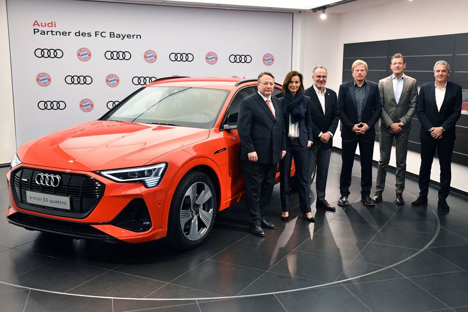 Vorstandsmitglieder des FC Bayern und Audi neben einem orangenen Audi e-tron