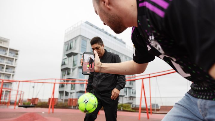 Two men playing soccer and one filming with cell phone